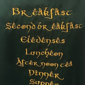 May include: A green fabric kitchen towel with gold embroidery that reads "Breakfast", "Second Breakfast", "Elevenses", "Lunch", "Afternoon Tea", "Dinner", and "Supper".