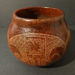 May include: A brown ceramic pot with a geometric design carved into the surface. The design is in a lighter brown color and features a star-like shape in the center.