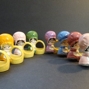 May include: Set of ten ceramic napkin rings featuring a girl in a bonnet and dress. Each ring is a different color: green, yellow, pink, green, pink, blue, red, pink, and yellow.
