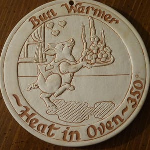 May include: A round, off-white ceramic bun warmer featuring an embossed illustration of a pig carrying a tray of buns. The text "Bun Warmer" is at the top, and "Heat in Oven ~350°" is at the bottom. There is a small hole at the top edge.