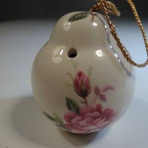 May include: A white ceramic ornament shaped like a pear with a pink and green floral design. The ornament has a gold cord attached to the top for hanging.