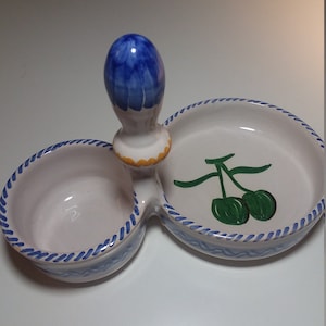 May include: A white ceramic double dish with a blue and yellow handle. The dish has a blue and white rope design around the edge and a green olive design on one side.