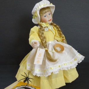 May include: A porcelain doll wearing a yellow and white gingham dress with a white apron and a white bonnet. The doll has blonde hair in braids and blue eyes. The doll is holding a small bowl in her hand.