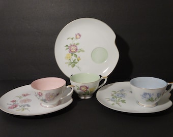 May include: Three floral teacups and saucers with matching plates. The teacups are pink, green, and blue with floral designs. The plates are white with floral designs.