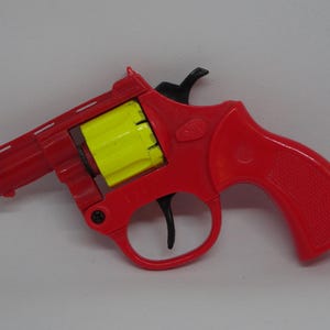 May include: A red toy revolver with a yellow cylinder. The toy gun has a black trigger and hammer, and a textured grip. The toy is made of plastic and is designed to look like a real firearm.