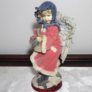 May include: A decorative angel figurine, featuring a red robe with white trim and a blue hood. The angel holds a basket of flowers and has large, detailed wings. The base is a dark red color.