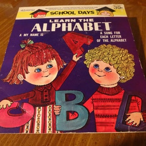 May include: A vintage record album cover featuring two children, a girl with red hair and a boy with blonde hair, holding colorful letters. The album is titled "Learn the Alphabet" and features the text "A song for each letter of the alphabet".