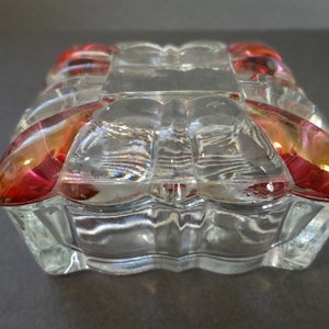 May include: A clear glass trinket box with a square shape and a decorative design. The box has a red and pink colored glass trim around the edges.
