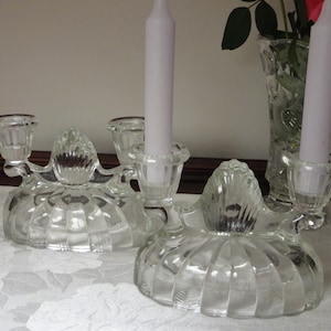 May include: Two clear glass candle holders with a three-candle design. Each holder has a scalloped base and a central decorative element. White candles are inserted in the holders. A vase with pink roses is in the background.