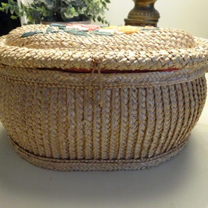 May include: An oval-shaped woven straw basket with a lid featuring floral embroidery in pink, yellow, and green. The basket has a braided texture, a small loop closure on the front, and two side handles made of the same woven straw material.