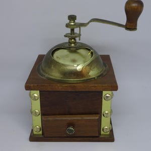 May include: A vintage coffee grinder with a wooden base and a brass top and handle. The grinder has a small drawer and brass accents. The wooden handle is a dark brown color.