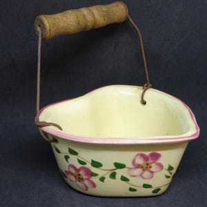 May include: A vintage, cream-colored ceramic basket with a pink rim and floral design. The basket has a wooden handle and metal wire supports. The flowers are pink with green leaves. A charming decorative piece.