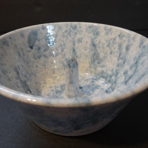 May include: A white ceramic ring holder with a blue and white speckled glaze. The holder has a raised center post for holding a ring.