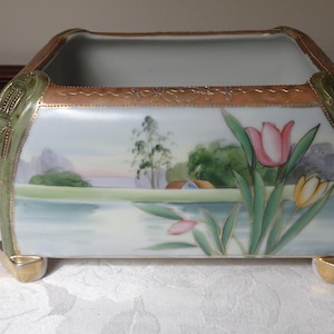 May include: A rectangular porcelain planter featuring a hand-painted landscape scene with a lake, trees, and a small house. The piece is decorated with pink and yellow tulips, gold-trimmed edges, green corner accents, and four gold-toned feet.