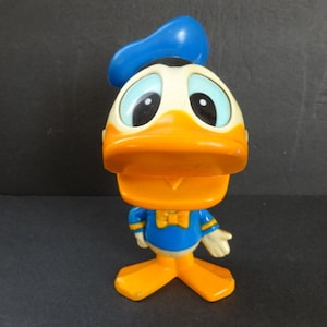 May include: A vintage plastic Donald Duck toy with a blue sailor hat, yellow beak, and orange feet. The toy has a blue and yellow sailor shirt and a bow tie.