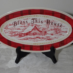 May include: Oval serving platter with a red plaid border and a central design featuring a red and white winter scene with a barn. The words "Bless This House" are written across the top.