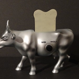 May include: A silver cow shaped toaster with a slice of bread in the slot. The toaster has a dial with the words "skim" and "whole" on it.