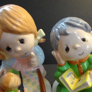 May include: Two ceramic figurines of children. A girl with brown hair is holding a baby doll and a quilt. A boy with gray hair is sitting and reading a book.