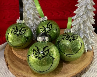 May include: Four green glitter Christmas baubles with a black Grinch face design. The baubles are on a wooden slice.
