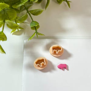 May include: Two beige cookie cutters with a bison shape and a small pink bison figurine on a white surface. Green leafy plant in the upper left corner. The cookie cutters are round with a raised edge.