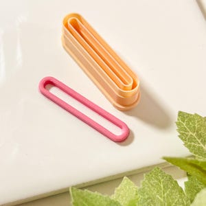 May include: A set of three plastic cookie cutters in the shape of an oval. The cutters are nested inside each other, with the largest cutter being a light orange colour, the middle cutter being a darker orange colour, and the smallest cutter being a pink colour.