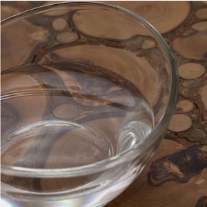 Puede incluir: Un cuenco de vidrio transparente, parcialmente lleno, se encuentra sobre una superficie de madera. La forma curva del cuenco refleja la luz, y la veta de la madera de la superficie es visible. La madera tiene un patrón natural y orgánico.