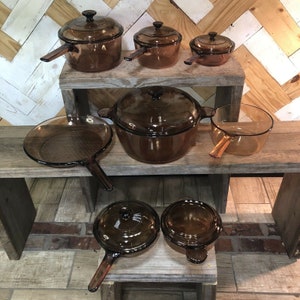 May include: A collection of amber-colored glass cookware including various pots, pans, and lids. The set features multiple saucepans with long handles, a large lidded casserole dish, a skillet, and smaller lidded vessels, all made of translucent brown glass.