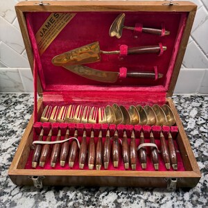 May include: A flatware set with gold-toned metal heads and dark brown wooden handles, housed in a wooden box with a red velvet interior. The set includes forks, spoons, and serving utensils. A label in the box reads JAMES.
