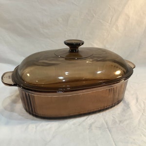 May include: An oval-shaped amber-coloured glass casserole dish with a matching lid. The dish features vertical ribbed detailing along the sides and integrated handles on both ends. The lid includes a central knob handle for easy removal.