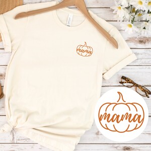 Op de afbeelding: Een wit T-shirt met een oranje pompoenontwerp met de tekst "mama" erop. Het shirt hangt aan een hanger en er is een aparte afbeelding van het pompoenontwerp op een witte cirkel.