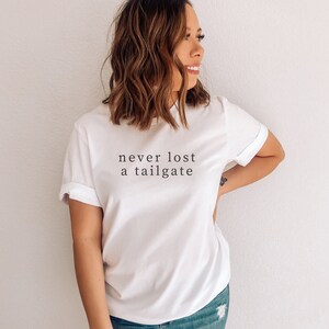 May include: White t-shirt with rolled-up sleeves featuring the text "never lost a tailgate" in a simple, black font. The shirt is made of a soft material and is suitable for casual wear.