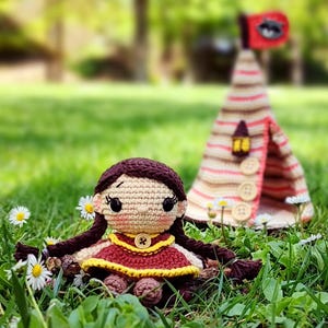 Puede incluir: Una muñeca de ganchillo hecha a mano con cabello castaño y un vestido rojo está sentada en un campo cubierto de hierba con margaritas. Una tienda de campaña de ganchillo a rayas con una bandera y una pequeña linterna está en el fondo.