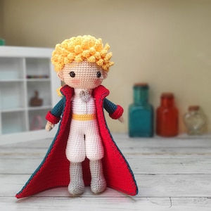 Puede incluir: Una muñeca de crochet del Principito, con una chaqueta roja y azul y una faja amarilla. La muñeca tiene el pelo rubio y una expresión dulce.