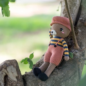 May include: A brown crocheted teddy bear wearing a pink and yellow striped shirt and a brown hat sits on a tree branch. The bear has a brown nose and black eyes.