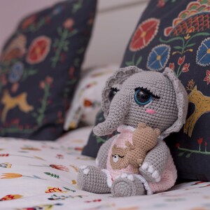 May include: A gray crocheted elephant plush toy with large blue eyes, holding a smaller brown crocheted bear plush toy. The elephant is wearing a pink dress with white lace trim. The plush toys are sitting on a white bedspread with a floral pattern.