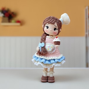 Pode incluir: Uma boneca de crochê com cabelo castanho em uma trança lateral, vestindo um vestido rosa com saia de babados azul, um avental branco e um chapéu de chef. A boneca segura um pequeno cupcake de crochê e possui detalhes faciais bordados.