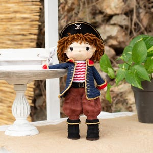 Puede incluir: Una muñeca de crochet que lleva una camisa a rayas azul y roja, pantalones marrones y un sombrero de pirata negro con un diseño de calavera y huesos cruzados. La muñeca está de pie sobre una superficie blanca con una bandeja de madera y una planta en maceta detrás.