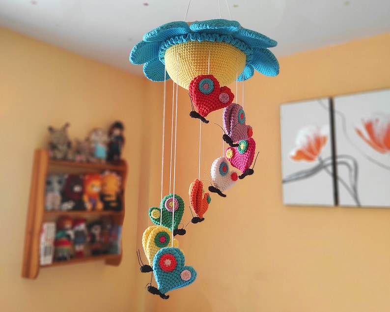 May include: A colourful crocheted baby mobile with a flower-shaped top and hanging butterflies. The butterflies are made of different colours of yarn and have a heart shape.