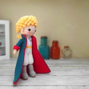 May include: A crocheted doll of The Little Prince wearing a blue and red cape. The doll has blonde hair and a white shirt.