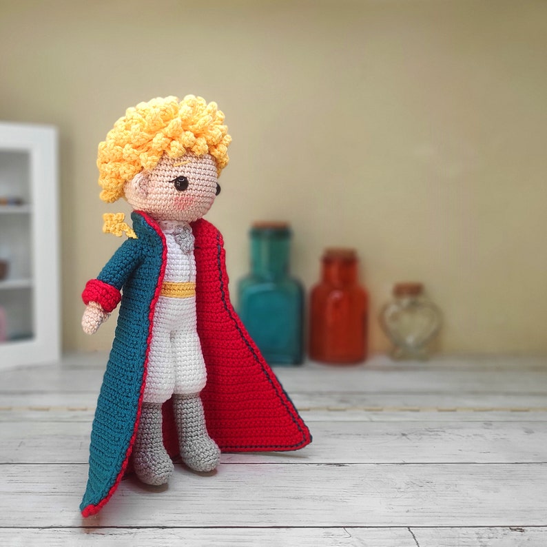 May include: A crocheted doll of The Little Prince wearing a blue and red cape. The doll has blonde hair and a white shirt.