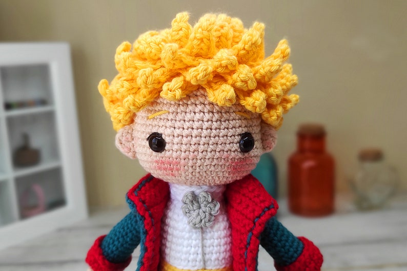 May include: A crocheted doll of The Little Prince, with a yellow hair, red and blue jacket, and a white shirt with a gray flower.