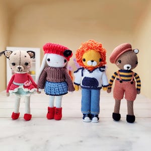 Puede incluir: Cuatro animales de peluche de crochet: un perro con manchas rojas y blancas, un conejo blanco con un sombrero rojo, un león con un suéter azul y blanco y un oso marrón con un sombrero rojo y un mono.
