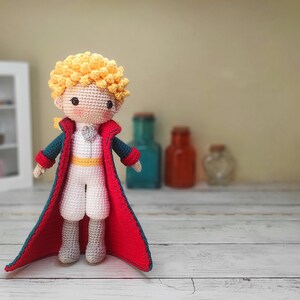 May include: A crocheted doll of the Little Prince wearing a blue and red jacket and a white shirt. The doll has blonde hair and a sweet expression.