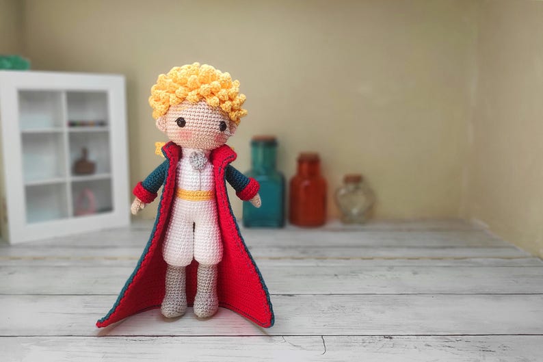 May include: A crocheted doll of the Little Prince wearing a blue and red jacket and a white shirt. The doll has blonde hair and a sweet expression.