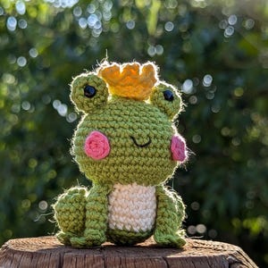 May include: A handmade crochet frog toy, wearing a yellow crown, with pink cheeks and black eyes. The frog is green with a white belly. It is sitting on a wooden surface, with a blurred green background.