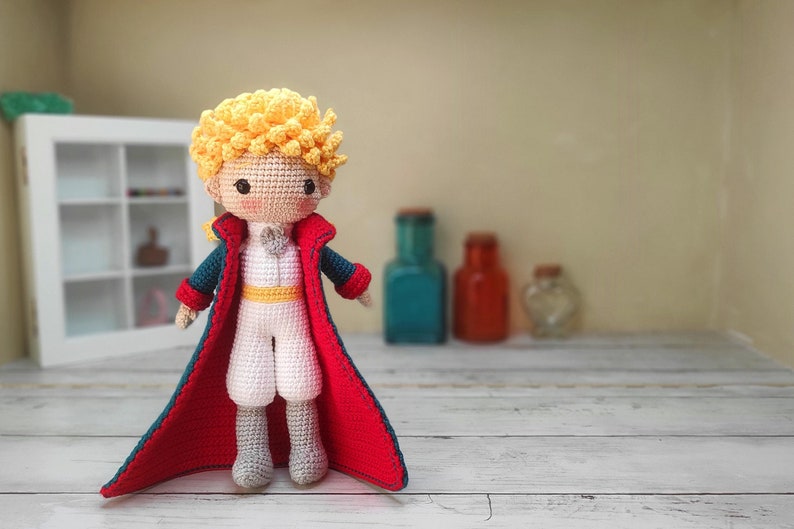 Amigurumi del Principito: muñeco tejido a mano con cabello rubio rizado, traje blanco y capa roja. De pie sobre una superficie de madera clara, con frascos decorativos en tonos azul y ámbar al fondo. Ideal para fans de este personaje clásico.