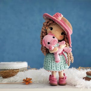 Puede incluir: Una muñeca de crochet con un sombrero rosa y marrón sostiene un cerdo rosa de crochet. La muñeca lleva un vestido azul claro y zapatos rosas. La muñeca está sentada sobre una superficie blanca y esponjosa.