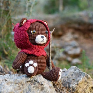 May include: A brown crocheted teddy bear wearing a red crocheted hood. The bear is sitting on a rock with a small stick in its hand.