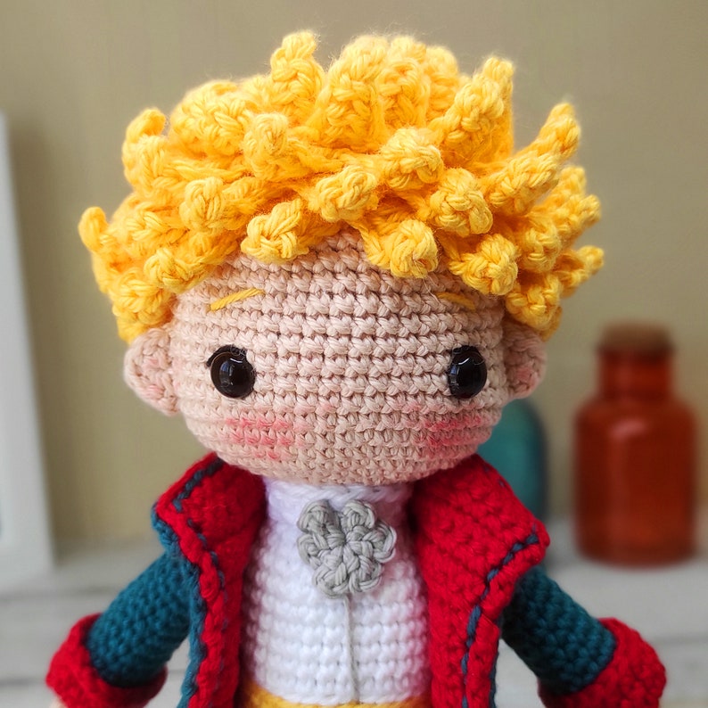 May include: A crocheted amigurumi doll of The Little Prince, featuring a yellow hair, a red and blue jacket, and a white shirt with a gray flower.