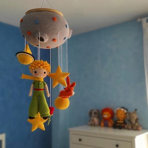 Può includere: Un mobile a crochet con un pianeta grigio, stelle gialle, una volpe e una figura del Piccolo Principe con abiti verdi e gialli.